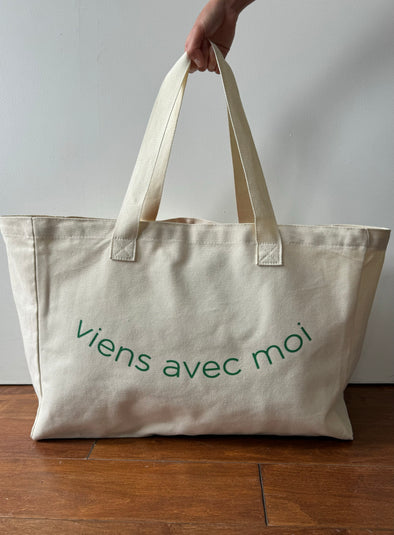 Oversized canvas tote in a natural cream colour. It has green print which reads viens avec moi in the shape of a smile.