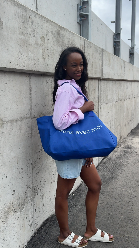 Oversized canvas tote bag by viens avec moi in royal blue, with white font print on the bag that reads viens avec moi in the shape of a smile.