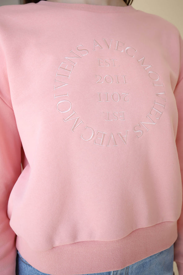 Closeup of the pink crewneck sweater with embroidered 'Viens Avec Moi' logo design on the front made from eco-friendly fabrics and designed in Canada.